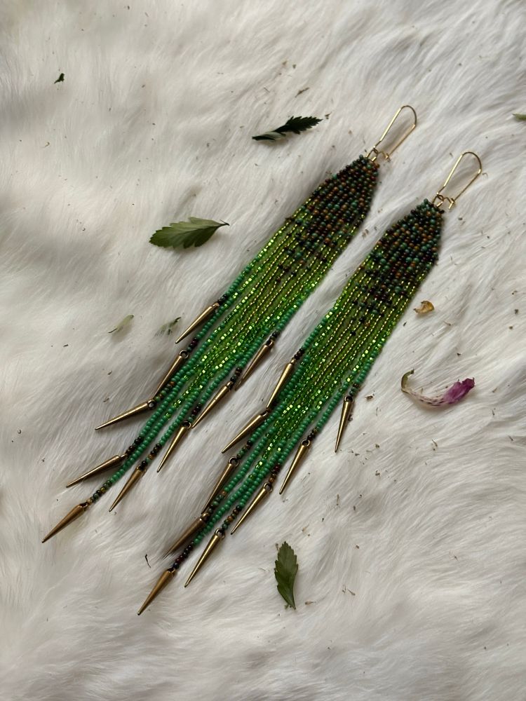 A green gradient pair of fringe earrings with bronze spikes at the ends. On a white rabbit fur backdrop with leaves & plant dusk scattered around. 