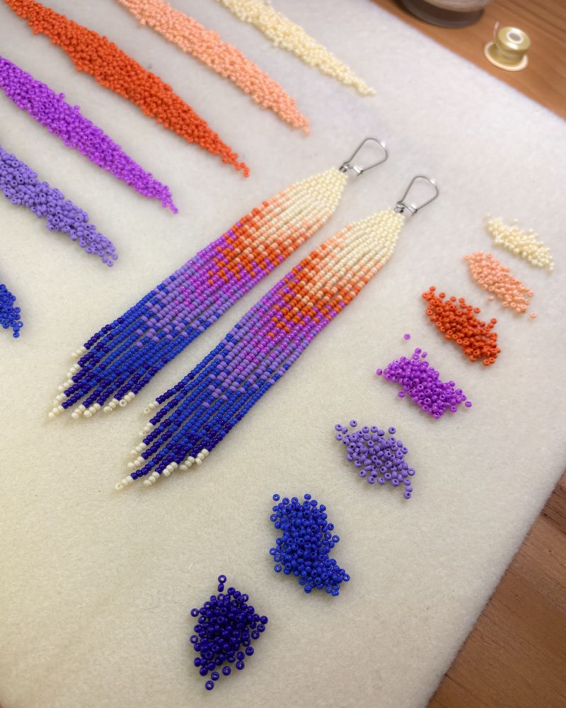 Sunset fringe earrings, a blend of cream, peach, orange, purple, blue & dark blue. photo is taken of a bead matte with all the bead colours of the earrinngs laid out. on the right in little circles. on the left in little lines.