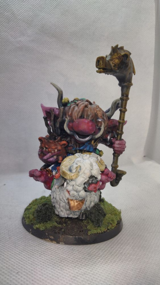 The Highland Goblin miniature from Rogue Hobbies. This is the mounted variant from the front. It has reddish brown hair, pink-purple skin, a big magenta nose with a silver nose ring. It is wearing blue tunic (?) and holding a red creature that resembles bagpipes. In its other hand its holding a large wooden staff topped with what looks like a razorback pig head carving. They are standing on a bass covered in green grass.  They are riding on a big woolly sheep with a bell around its neck.