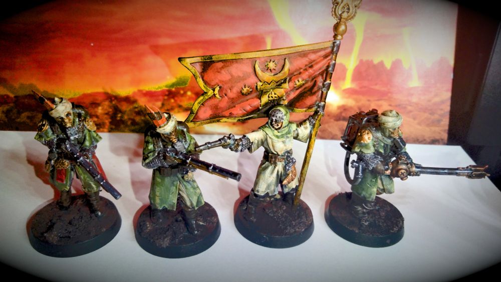 The four Azeb miniatures from the Trench Crusade line. The 2 on the left and the one on the right are wearing military green overcoats and cream head coverings. The 2 on the left are wielding rifles and the 1 on the right is wielding an exotic flamethrower. They are also wearing golden gas masks. The 2nd from the right is wearing cream robes with a military green head covering. She is wearing goggles. She is wielding an archaic pistol and holding a red flag with star and crescent moon imagery. All are wearing chain mail under their clothes. All have muddy bases and are standing in front of an image of a fiery landscape.