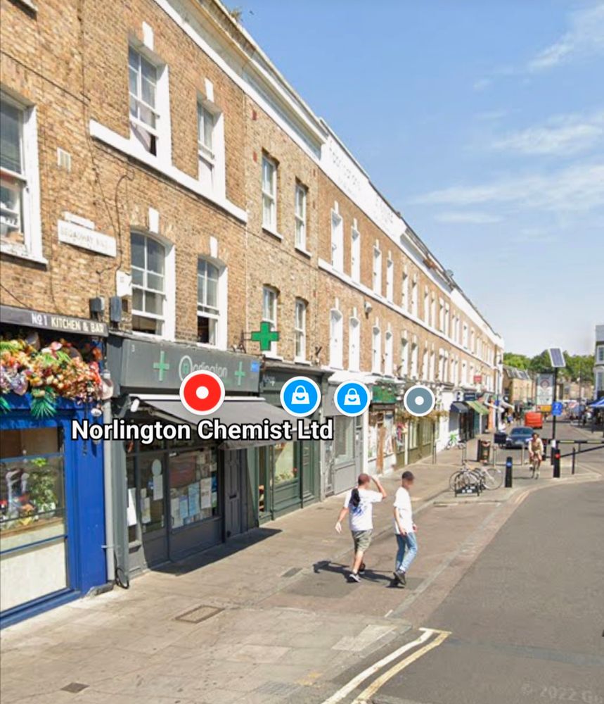 Street view of independent shops on a local high (main) street with two people walking away, and further down the road, another person on a bicycle is cycling towards the viewer.