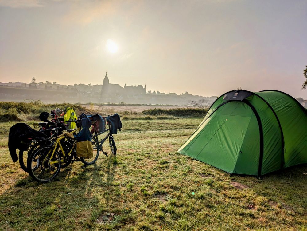 Four bicycles standing next to a green tent in the early morning light. In the background is a silhouette of a town.