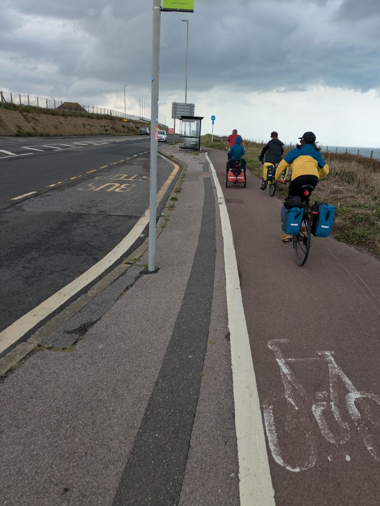Cyclists riding on a shared path with painted bike in the right hand side of the path and a bus stop shelter on the left, beside a busy road.