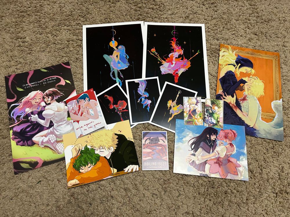 Pile of merch containing the following: one mizisua (alien stage) poster, one mizisua fan comic, one bakudeku (mha) mini poster, one Madohomu (Madoka Magica) mini poster, two Madoka Magica posters (one of Homura, one of Madoka), three Madoka Magica postcards in the same style (one of Kyoko, one of Sayaka, one of Mami), one Boiling Isles (the owl house) postcard, two mha gold foil character cards (one of Bakugou, one of Deku), and a farcille (delicious in dungeon) poster.