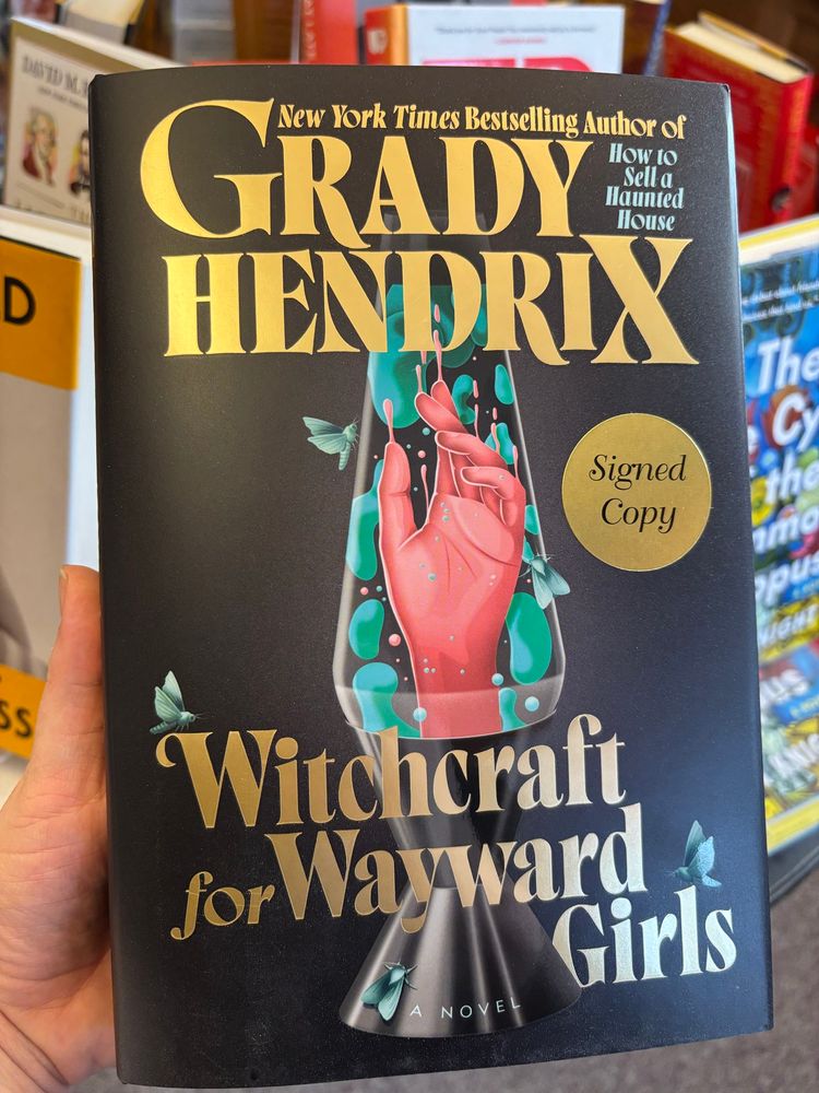 A black book with gold lettering. The title is Witchcraft for Wayward Girls: A Novel by Grady Hendrix. On the cover is the drawing of a lava lamp with teal green blobs and a florescent pink hand inside. Flys sit on the lava lamp and title. 