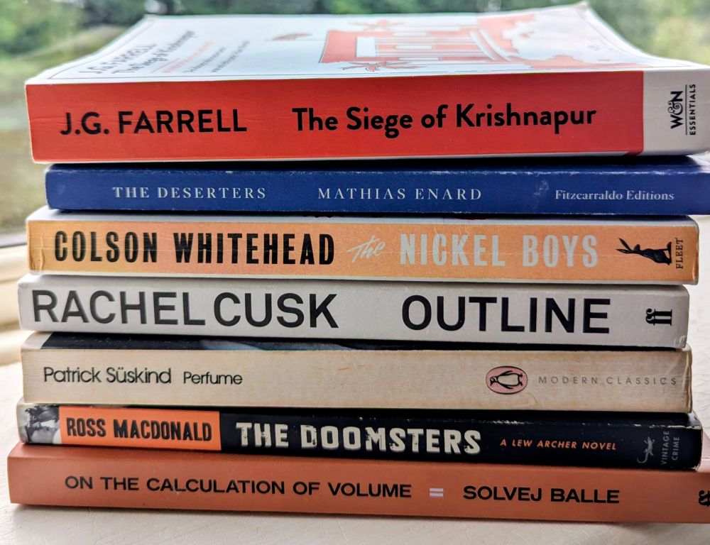 A pile of paperback books on a window ledge: The Siege of Krishnapur, The Deserters, The Nickel Boys, Outline, Perfume, The Doomsters, On the Calculation of Volume, Part Two. 