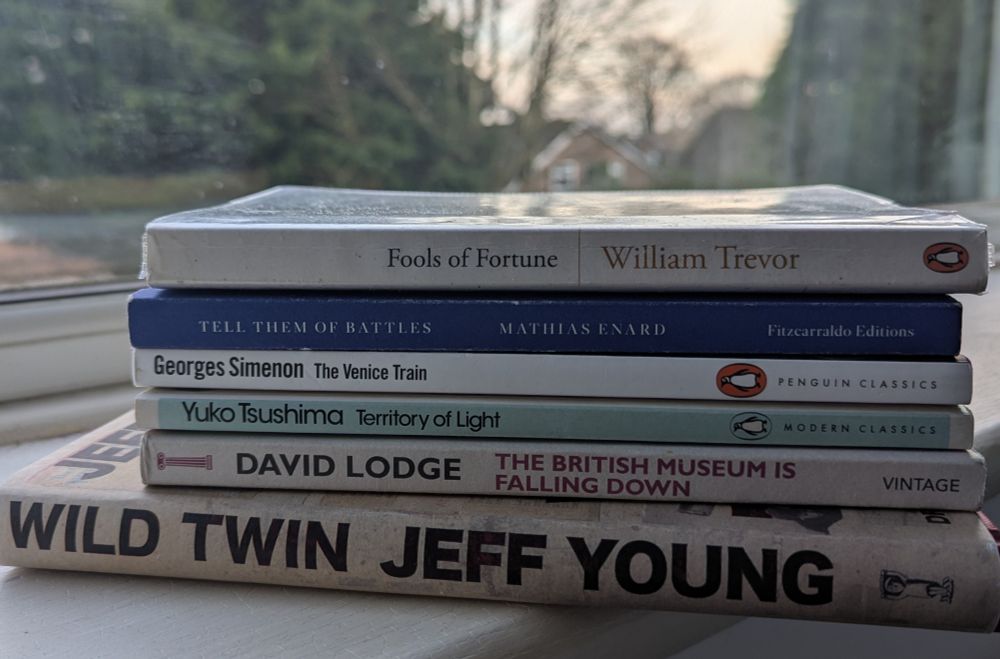 A pile of paperback books by a window: Fools of Fortune (William Trevor); Tell Them of Battles (Mathias Enard); The Venice Train (Georges Simenon); Territory of Light (Yuko Tsushima); The British Museum is Falling Down (David Lodge); Wild Twin (Jeff Young).