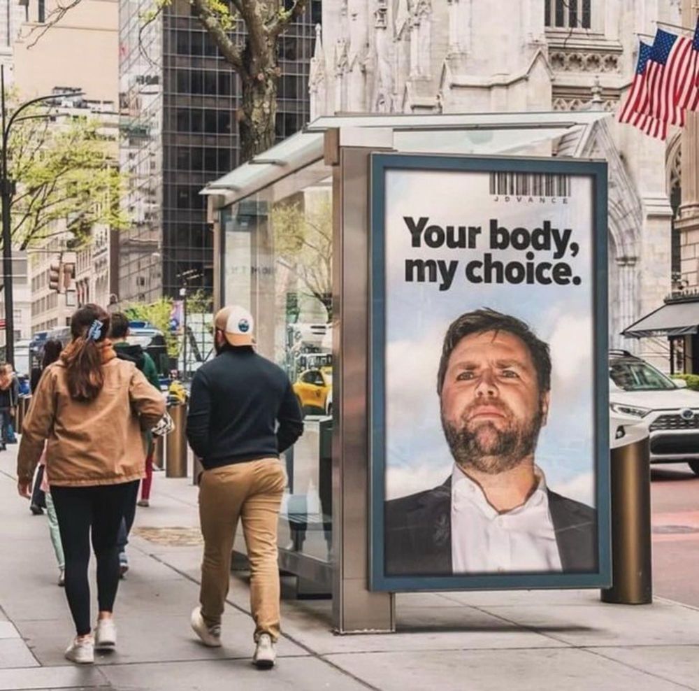 Photo of a city bus stop, a couple of people are walking past the bus stop. On the side of the bus stop is an ad that shows a head & shoulder shot of JD Vance. Above his head are the words "Your body, my choice".