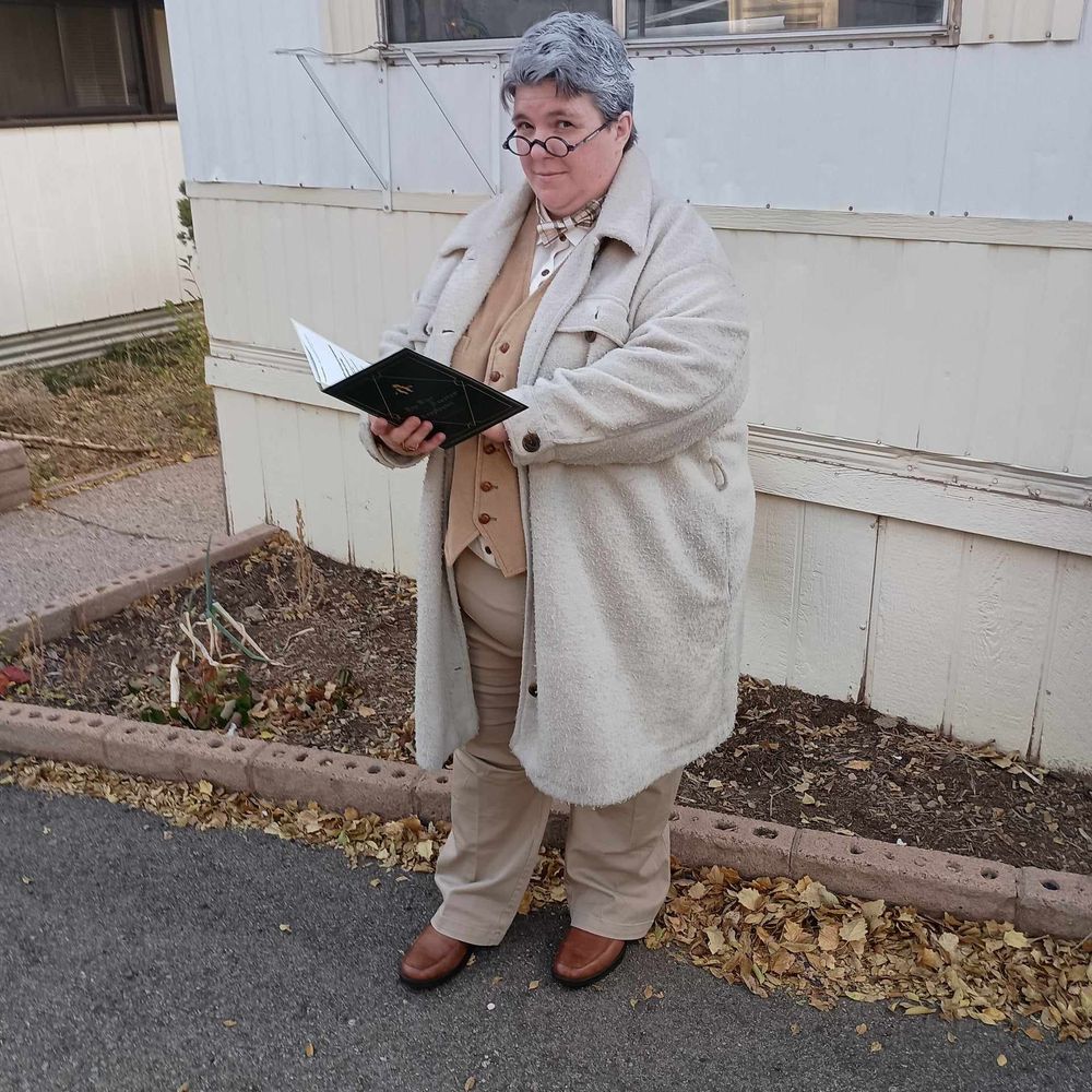 Cosplay of Aziraphale: fat woman with grey hair wearing small reading glasses, brown waistcoat, long cream jacket, light brown pants, and brown shoes, holding a green book and looking at the camera.