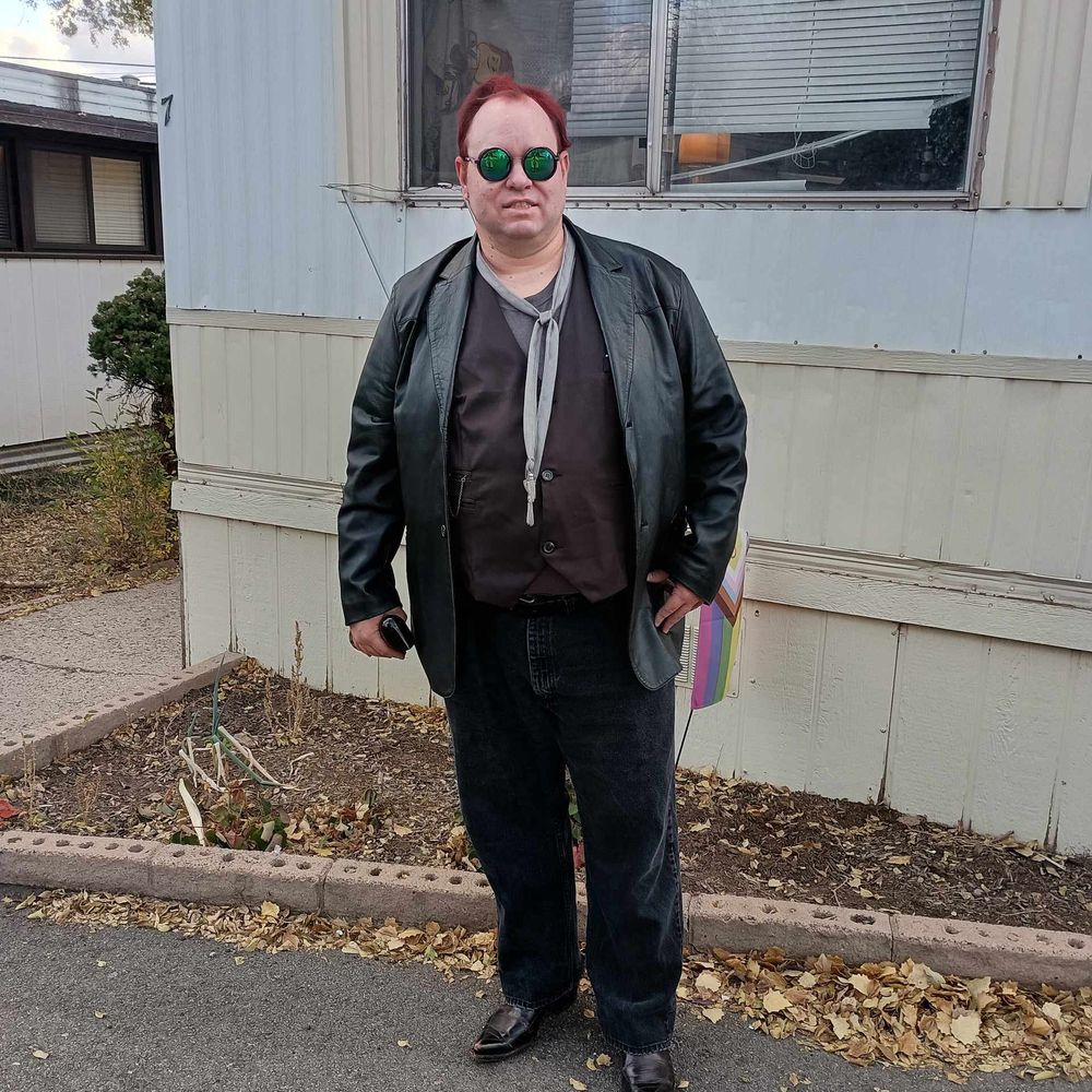 Cosplay of Crowley: man with red hair wearing reflective sunglasses, black leather jacket, loose silver necktie, black waistcoat, black jeans, and holding a spray bottle in front of a PRIDE flag and trailer.