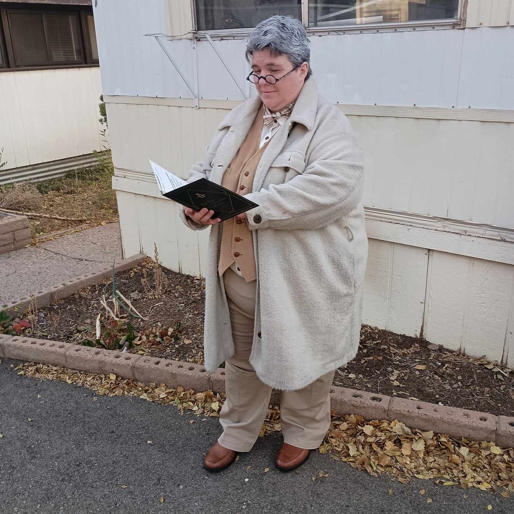 Cosplay of Aziraphale: fat woman with grey hair wearing small reading glasses, brown waistcoat, long cream jacket, light brown pants, and brown shoes, reading a green book in front of a trailer.