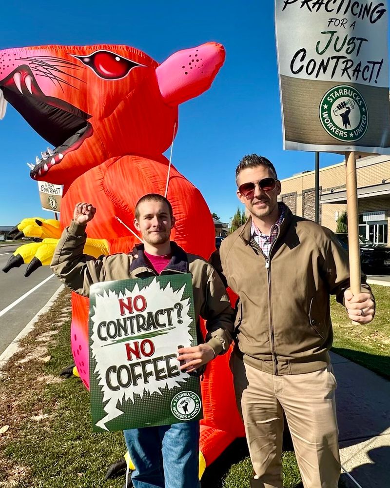 Adam (right) with SBWU supporter Max, holding a no contract, no coffee sign. Red inflatable rat (scabby) in the background 