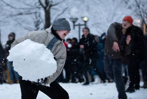 a kid holds a large heavy snowball that's almost too havy for him in both hands preparing to throw it at the other person in the snowball fight
