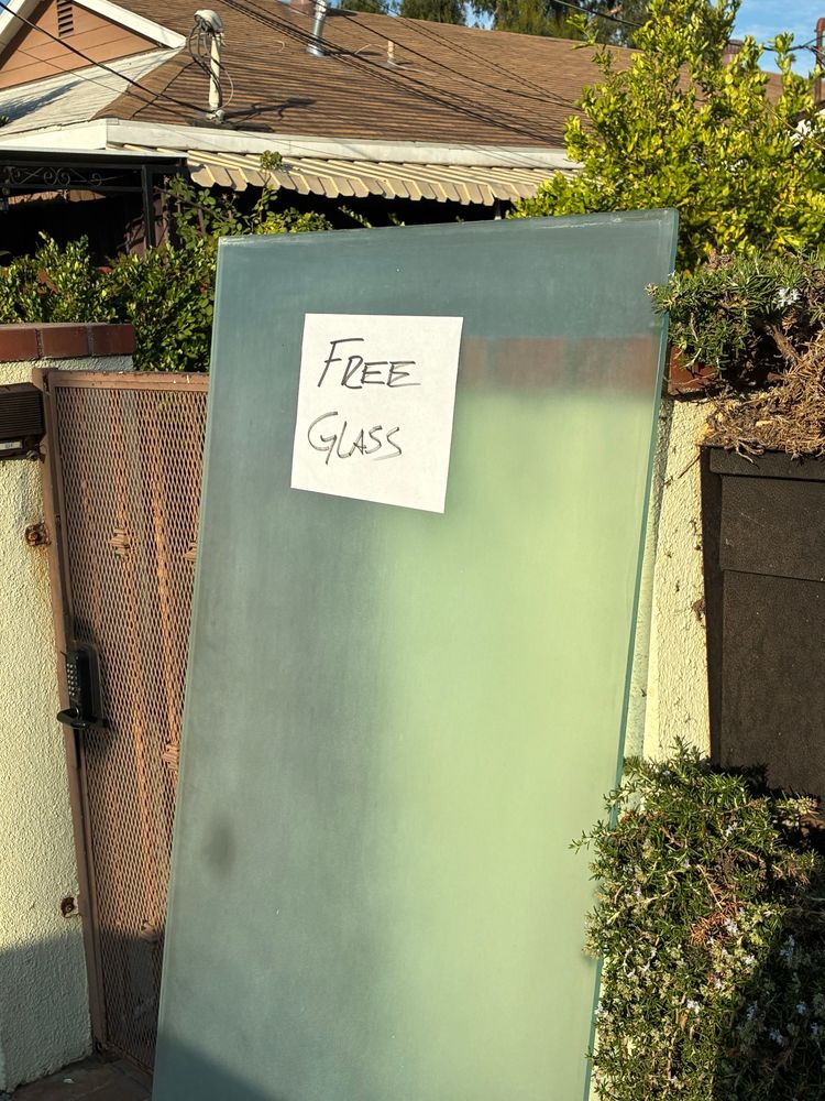 door-sized panel of frosted glass leaning against a home fence with a piece of paper taped to it that says “FREE GLASS”