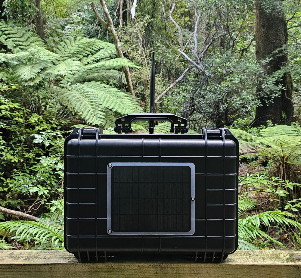A black rugged case with handle and solar panel affixed to front, antenna mounted vertically behind the handle. It sits atop a wooden rail, with native bush/forest behind it