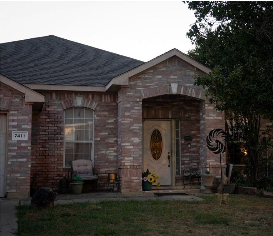 Brick house with an arched entryway at dusk. A round-top window on the left has a chair beneath it. Sunflower decor and potted plants by the door.
