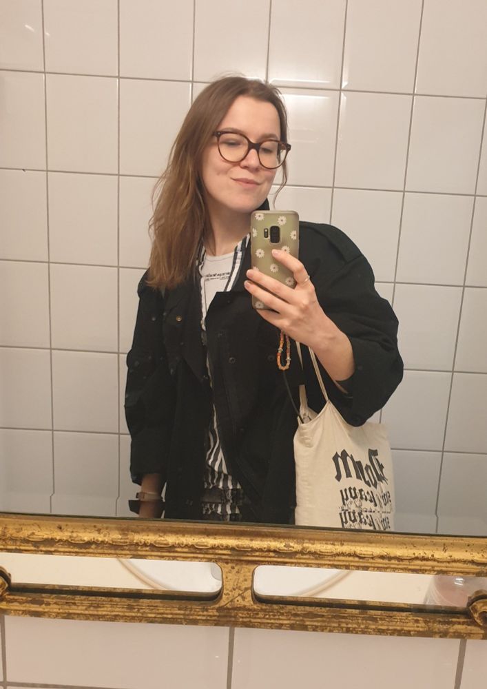 Spiegelselfie: ich mit langem braunen Haar, einem schwarzen Oversizemantel und einem Jutebeutel über der Schulter fotografiere mich selbst in einem Spiegel mit golgenem Rahmen