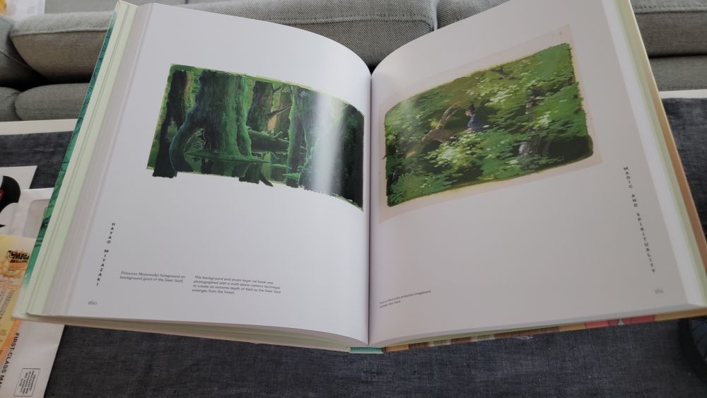 An open Hayao Miyazaki art book from the Academy Museum Press lies on a gray fabric surface.
Left page: A wide background painting from Princess Mononoke shows a dense, ancient forest. Massive moss-covered tree trunks rise vertically, their roots twisting over fallen logs and deep shadows. The palette is rich in greens, creating a sense of mystical depth and age. A caption beneath notes that the multilayered background was photographed using a multi-plane camera technique to achieve extreme depth of field.
Right page: Another production painting from Princess Mononoke depicts a lush forest clearing dappled with light. A small human figure rides a creature through the greenery, partially obscured by bright leaves and soft shadows. The image feels airy and alive, capturing the film’s blend of natural beauty and quiet spiritual energy. The page is labeled “Magic and Spirituality” along the right margin.