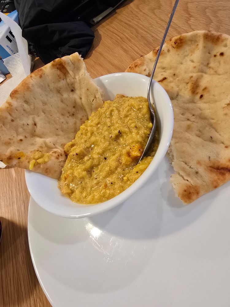 A white bowl of golden-yellow mulligatawny soup, thick and creamy with visible spices, sits on a white plate. A metal spoon rests in the bowl, and two pieces of garlic naan are propped on either side, slightly charred and folded over the rim. The setup is on a light wooden table with a few items in the background.