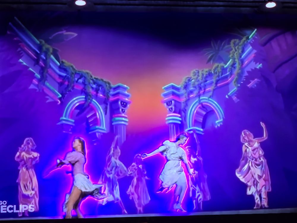 A vivid scene from Xanadu (1980) during the opening number “I’m Alive.” The screen shows a group of dancers bathed in glowing neon light against a surreal, painted backdrop of Greco-Roman arches. The arches are outlined in luminous blues and pinks, set against a twilight gradient sky of orange and purple. Two dancers in the foreground, wearing flowing pastel costumes, move energetically toward each other, while ethereal figures of other muses appear behind them as if emerging from the mural itself. The overall effect is dreamlike and electric, blending live action with glowing animation.
