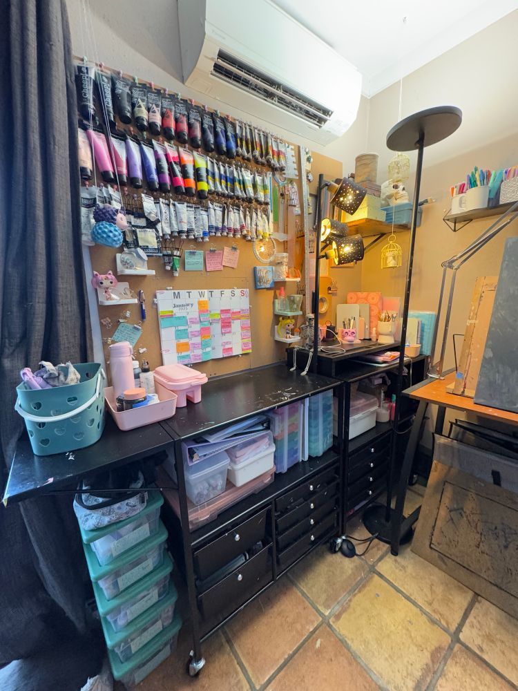 A at home art studio and gaming space. By the window curtain along the wall is a pair of metal carts with drawers laden with supplies. Above that is a wall of cork boards covered in paint and other items, along with a calendar covered in todo items. At the far side of the wall is several shelves covered in books and more materials. On the tile floors there are larger books in canvases  in bags. There are small completed art pieces scattered about. 

the adjacent wall has more shelves with markers in small cups. There is a desk with a drafting easel on it and a recycled canvas painted a deep grey