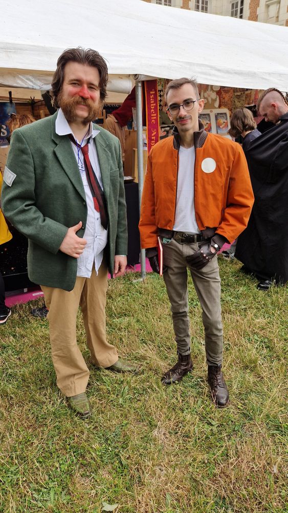 Picture of my boyfriend and I taken while cosplaying as the main characters from Disco Elysium, I am wearing a green vest with a red tie and a white shirt, beige pants, and pointy shoes that are dyed green. I have mid lenght hair that are brushed back and mutton chops, I also have red makeup on the nose and part of the cheeks to imitate a rosascea; my boyfriend is wearing kakhi slim pants, a pair of brown ankle boots, a white t-shirt, an orange vest with white patches, round glasses and dark red leather gloves, his hair are cut short and he has moustache stubble visible.
In the background you can see lots of tents with stands inside, and people walking around checking out the stands.
