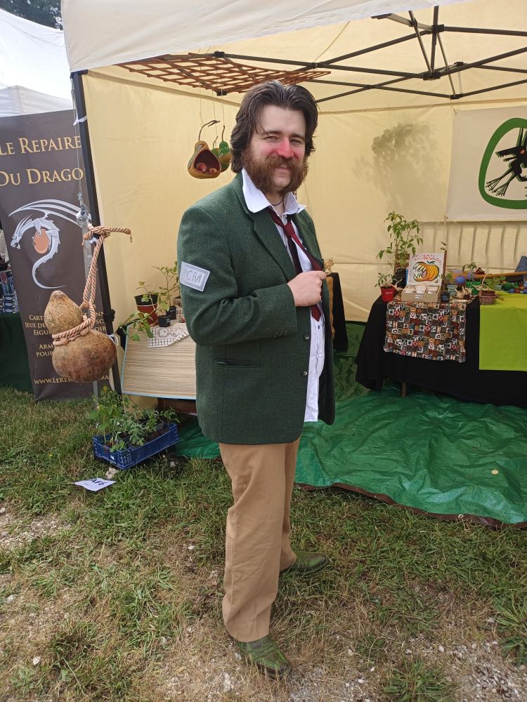 Picture of myself cosplaying Harry Dubois from Disco Elysium, I am wearing a green vest with a red tie and a white shirt, beige pants, and pointy shoes that are dyed green, I have mid lenght hair that are brushed back and mutton chops, I also have red makeup on the nose and part of the cheeks to imitate a rosascea. I am standing while holding my tie between my fingers.
In the background you can see a tent with a stand inside that has plants put on top.