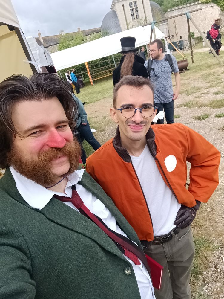 Picture of my boyfriend and myself taking a selfie while cosplaying as the main characters from Disco Elysium, I am wearing a green vest with a red tie and a white shirt, I have mid lenght hair that are brushed back and mutton chops, I also have red makeup on the nose and part of the cheeks to imitate a rosascea; my boyfriend is wearing kakhi slim pants, a white t-shirt, an orange vest with white patches, round glasses and dark red leather gloves, his hair are cut short and he has moustache stubble visible.
You can see tents with tables under planted in grass in the background, as well as a castle behind, with a cloudy sky.
This cosplay was described as "perfectly accurate" by attendees who recognized us during this convention.