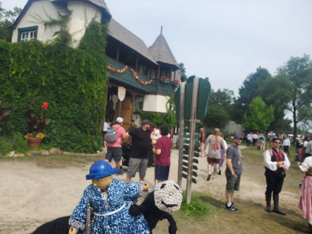 A wood doll in crochet dress and a crochet long doll in front of Ren Faire gates