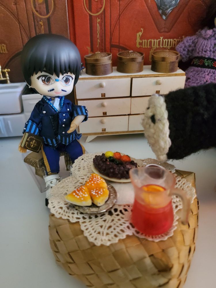 Martyn (a nendoroid doll in blue clothes with glasses, holding a book in one hand) and Dollmaker (a black crochet creature with a white face) around a table with tiny food on it. 