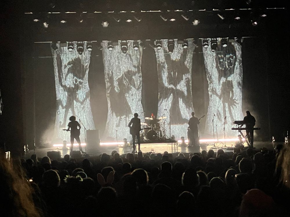 picture of the band Car Seat Headrest onstage. the stage is lit such that all five band members are still shrouded in shadow, and now there are four anthropomorphic cartoon figures visible on the stage backdrop.
