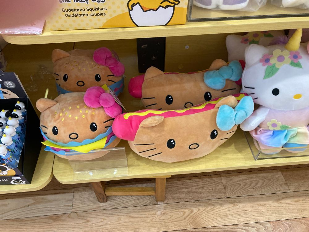 Photo of plushies sitting on a shelf. They are Hello Kitty as a hot dog in a bun and as a cheeseburger.