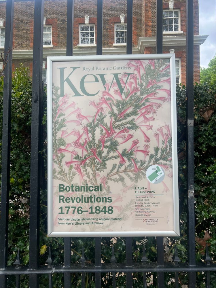 Sign attached to a fence on the outside of the Kew Gardens building. The sign, which has a large image of a green and red plant, describes an exhibition on the history of botany currently hosted inside Kew building.
