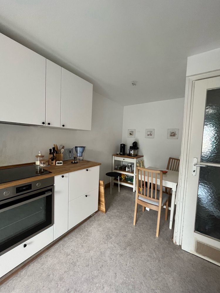 Links ein Stück der Küchenzeile, rechts der Tisch mit zwei Stühlen und im Hintergrund noch ein Küchenwagen in weiß ebenfalls mit eichefarbener Arbeitsplatte drauf