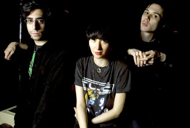 A photo of the rock band YeahYeahYeahs. They are looking up at a camera with drummer Brian Chase on the left, singer Karen O in the center and guitarist Nick Zimmer on the right. 