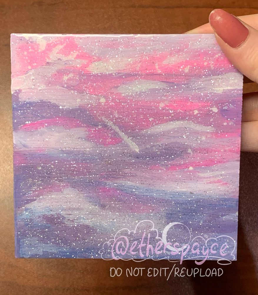 A picture of a coaster painted in soft pinks and lavender of a shooting star crossing through layers of clouds. A resin coat makes it appear glittery and shiny.