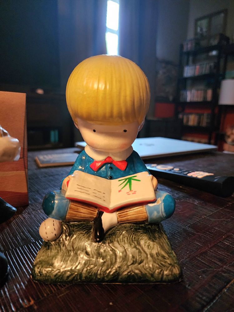 A ceramic figure of a little boy with blond hair, wearing a blue suit, sits on the grass, next to a baseball, reading a book with an image of a red flower.
