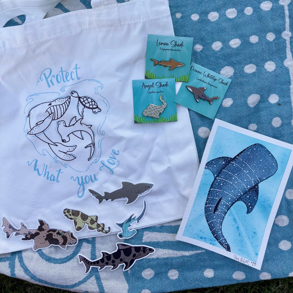 Image depicts 3 enamel pins (a lemon shark, an angel shark, and an oceanic whitetip shark), 5 stickers (a horn shark, a spotted wobbegong shark, a hammerhead shark, a tiger shark, and a leopard shark), a tote bag with the illustrated image of a whale, a hammerhead shark, and a sea turtle arranged in the shape of a heart with the words “protect what you love” around it, and a small watercolor painting of a whale shark viewed from above, all spread out on a blue and white beach towel. Everything in the photo is included in the giveaway on my Instagram page except for the towel. 