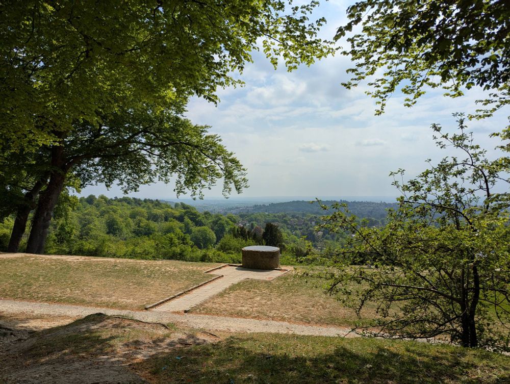 A hilltop view across a valley, framed by tree canopy, center frame is a large stone block with inscriptions of local places 