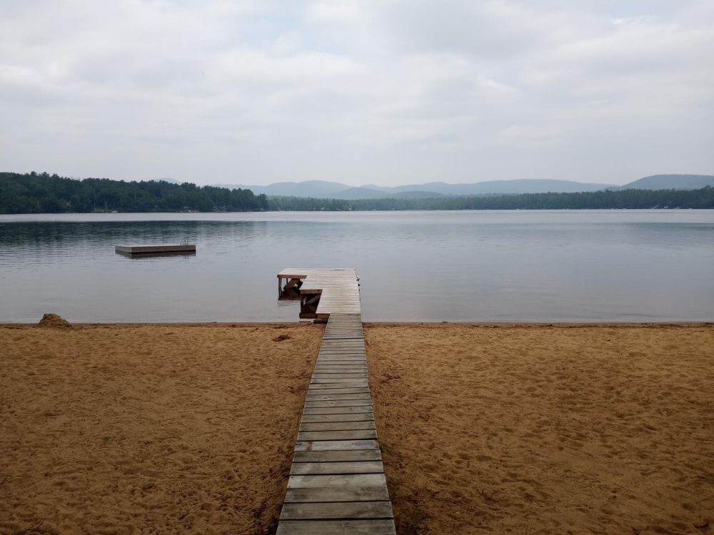 A photograph of a lake and sky with sand in the foreground. There is a wooden pier/dock intersecting verically and a floating platform to the left. A thin line of trees and mountains separates the sky and lake.