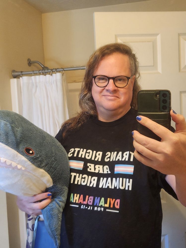 Talia looking at the camera, wearing a Dylan Blaha pride t shirt that says "trans rights are human rights" She is holding a blahaj under her left arm.