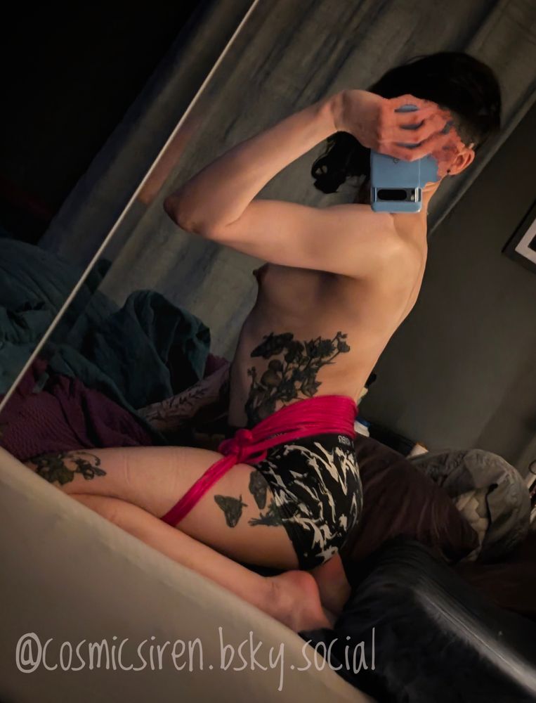The non-binary wild magic condensate is on their knees in a bed. Their thighs are bound to each other behind the back holding them in place solely because the butt is too round. They are doing their best to sit up but it's difficult. Their tattooed arm is being used to help hold them up and the other one is holding the phone taking this picture. A breast is visible, only partially obscured by the leanly toned arm. You can clearly make out the curve of their back and side tattoos visible as they waver ever so slightly at the tension forcing them back down. 