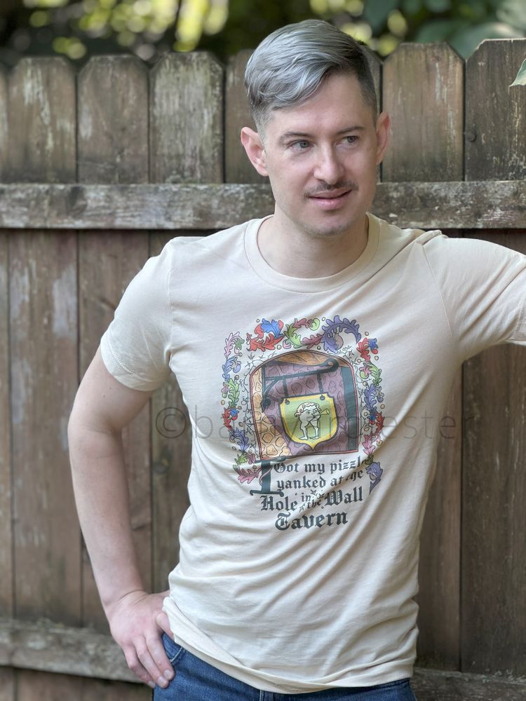 A model wearing an offwhite t-shirt with colorful medieval scrollwork. In the center of the artwork is a sign with a humanized ass tooting a horn. The medieval text reads "I got my pizzle yanked at the Hole in the Wall Tavern."