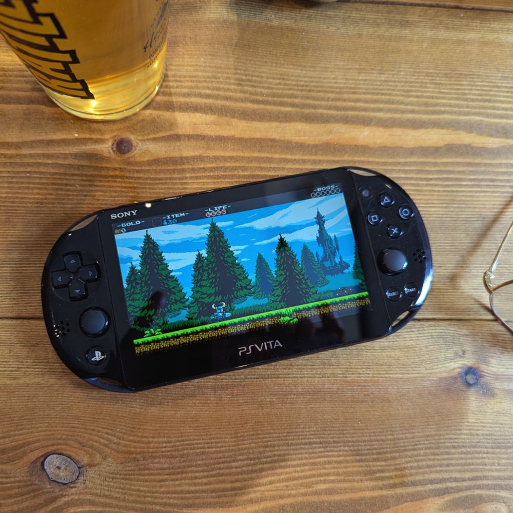 A PlayStation Vita playing Shovel Knight, lying on a wooden table next to a glass of cider and some spectacles.