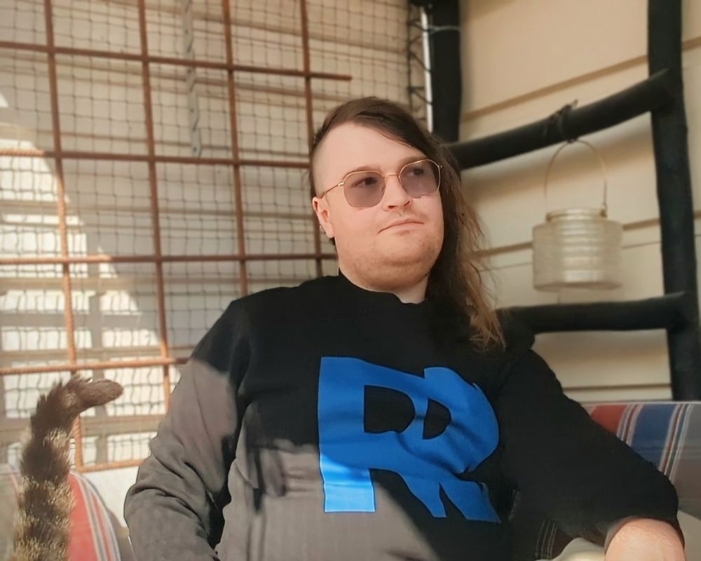 A picture of me wearing a black sweater with the logo of Remedy games printed on it in blue. A brown cat is just out of frame but the tail can be seen.
