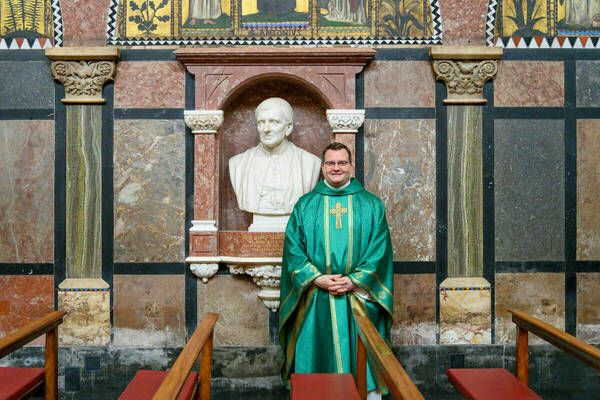 A person in green religious vestments smiling at the camera, standing in front of a marble wall with a bust of St. John Newman in the background, inside a richly decorated church.