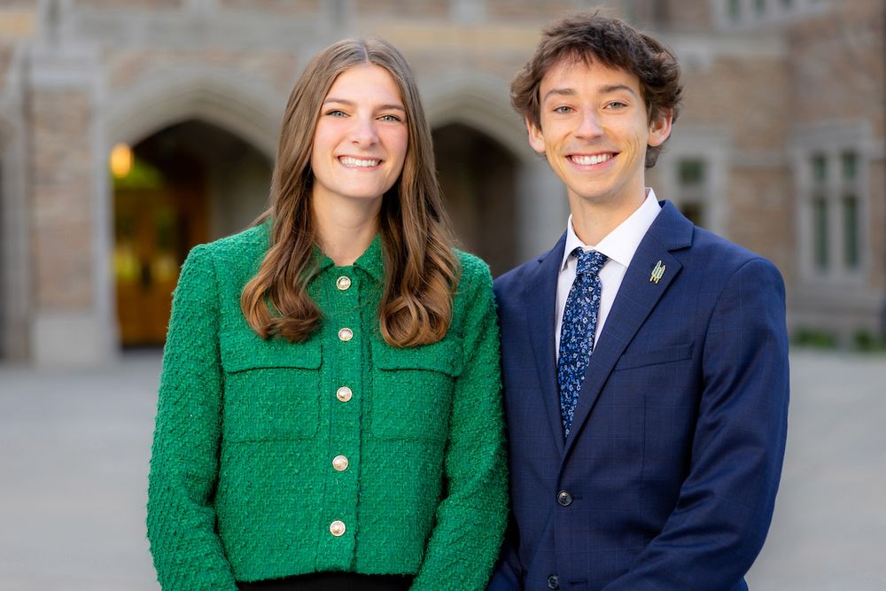 Two students smiling in front of a stone archway, one wearing a green jacket and the other in a blue suit with a lapel pin.
