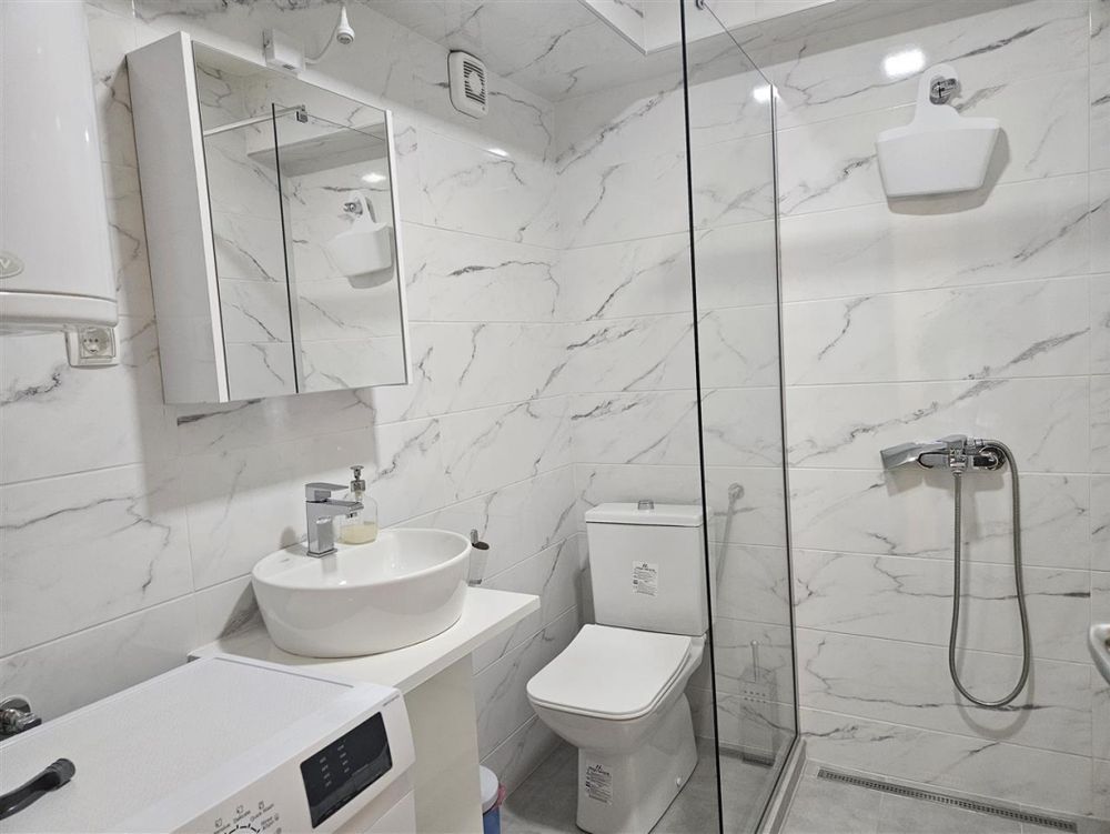 picture of a clean white bathroom in a third apartment. on the left is a washing machine and a sink. on the right is a shower stall. in the corner between the wall and the wall of the shower stall, directly beside the sink, is a boxed in toilet