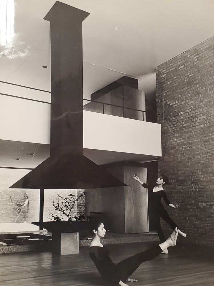 Photograph in black and white showing two dancers in leotards in simple dance positions, posing inside a modern home, in front of a suspended fire hood and double height loft