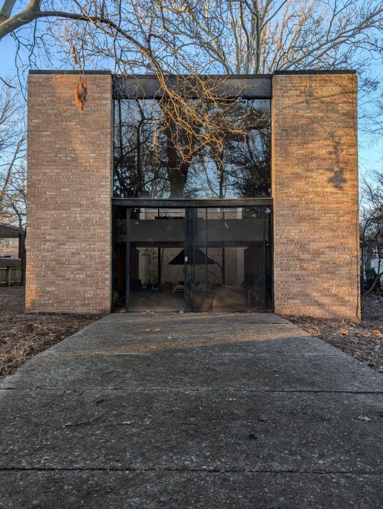 Photograph of the rear facade of a house showing a double height glass window and door at center with red brick corner piers and a pebbled concrete patio
